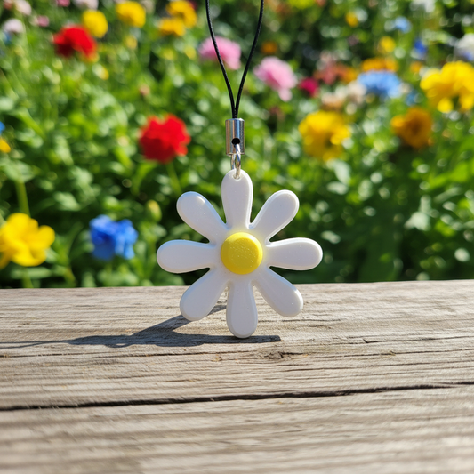 Brelok Stokrotka 3D w kolorze białym z żółtym środkiem na drewnianym tle, w tle kwiaty w różnych kolorach